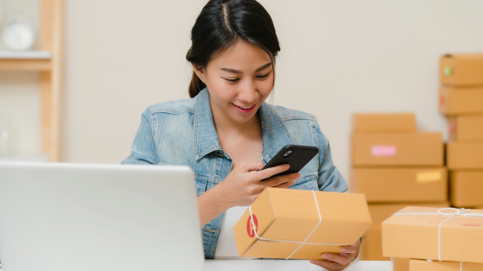 A small business owner checking a parcel against her phone beside a laptop and stacked cartons, representing ordering wholesale goods online from China