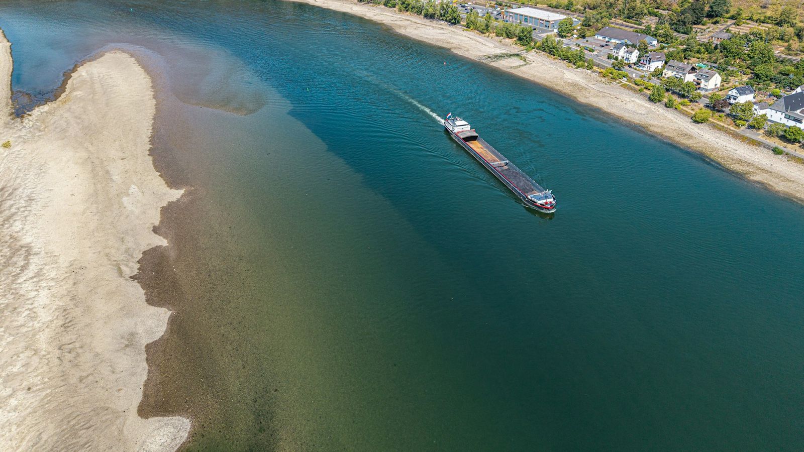Aerial view of a cargo barge navigating a narrow deep channel between wide exposed sandbanks on a river, representing the draft limits of an inland river port
