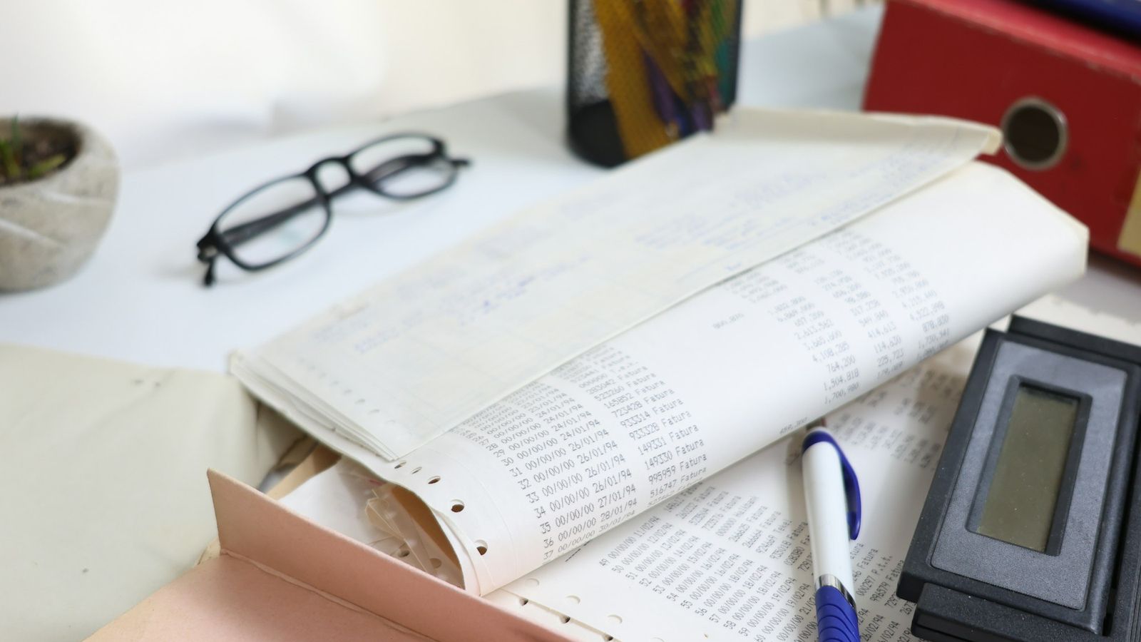 A desk with a long columnar printout of figures, reading glasses, a calculator and a ring binder, representing customs assessment paperwork
