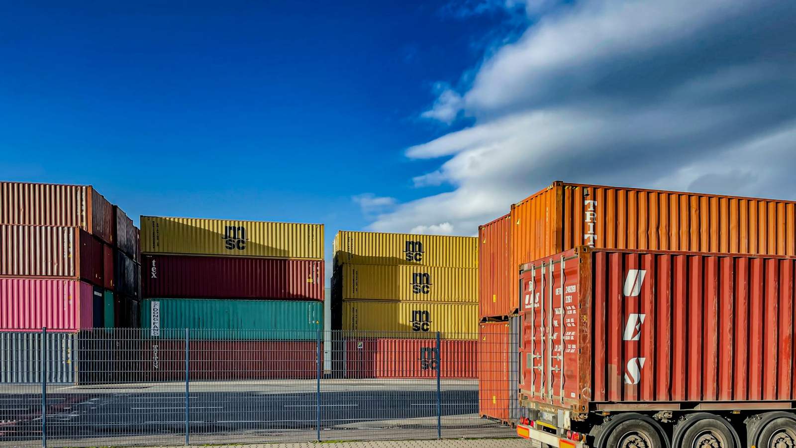 Stacked shipping containers behind a fence at a freight terminal, with one container loaded on a truck trailer, representing cargo waiting to be released