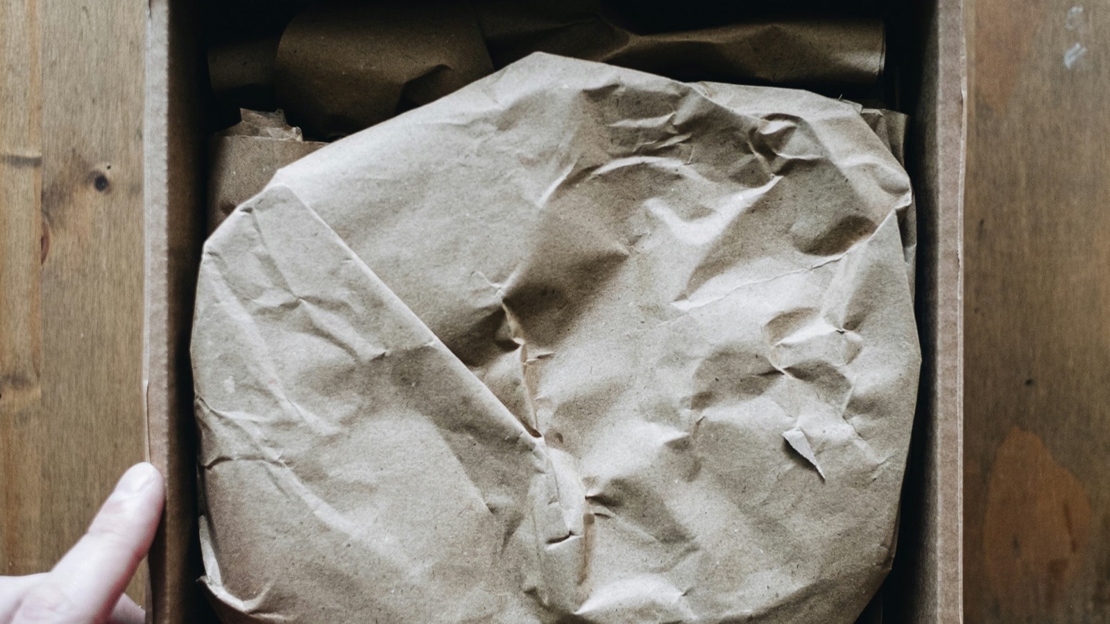 A hand opening a cardboard box to reveal an item wrapped in brown kraft paper, representing the arrival of a product sample from a supplier