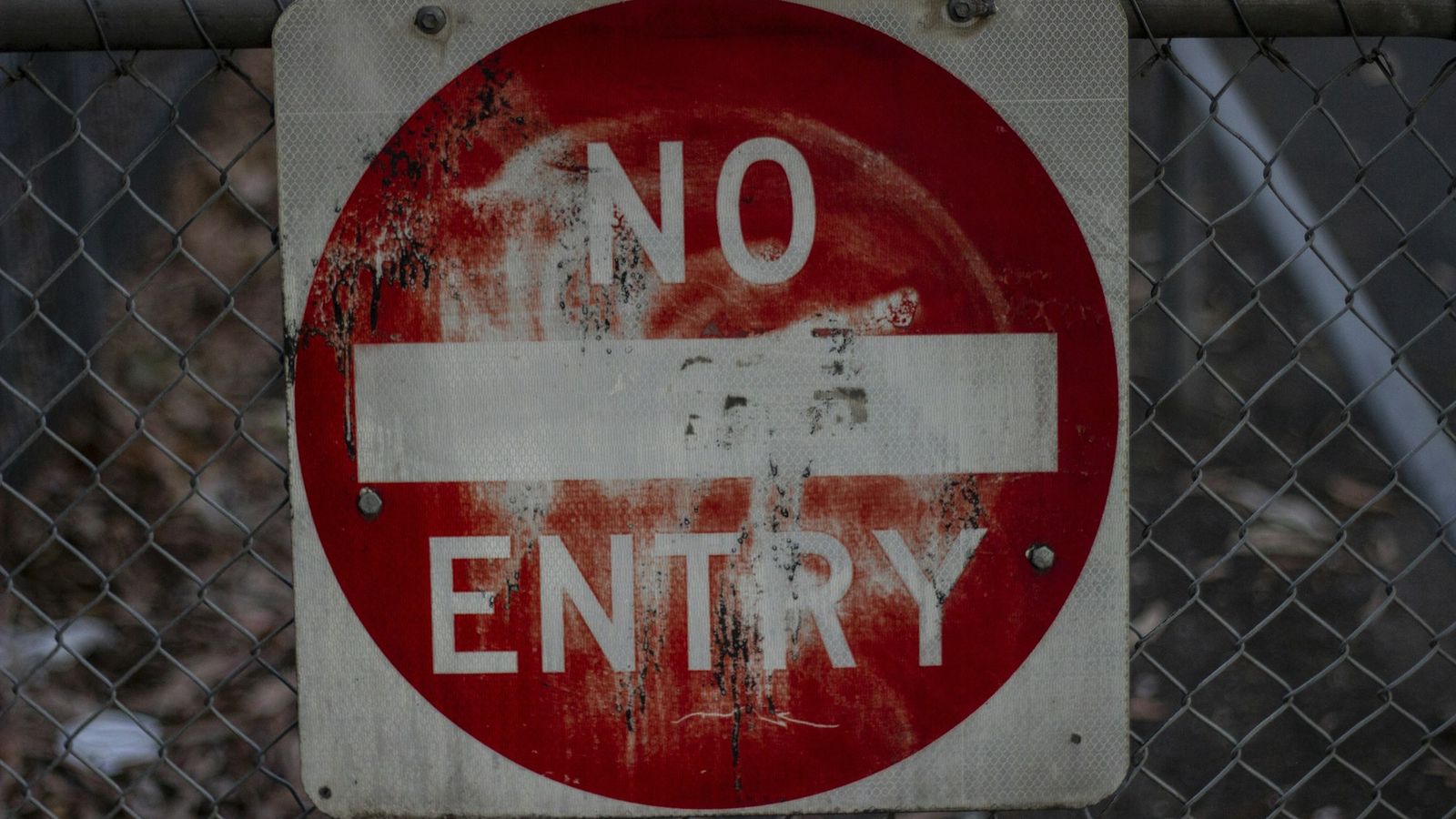 A weathered red and white NO ENTRY sign mounted on a chain link fence, representing goods that cannot be brought into the country